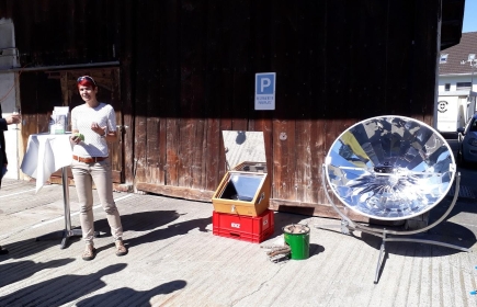 Petra Bolfing präsentiert drei Kocher, vlnr: Solarkocher, engergiespar Holzkocher, Parabolspiegelkocher